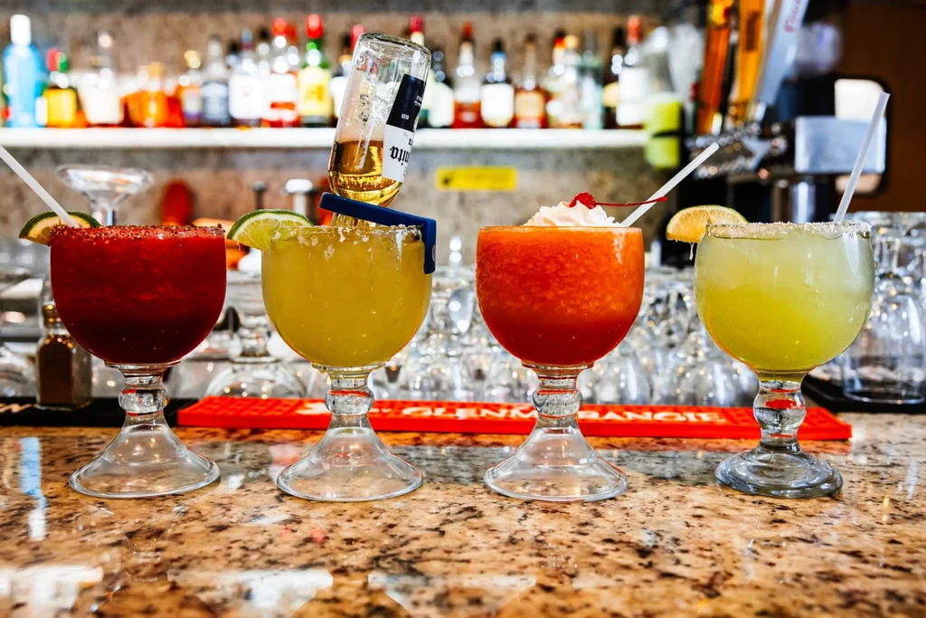 Four vibrant cocktails on a bar, one with an inverted Corona bottle.