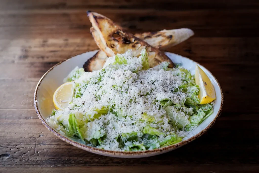 Close-up Caesar salad with Parmesan, bread, and lemon on dark wood.