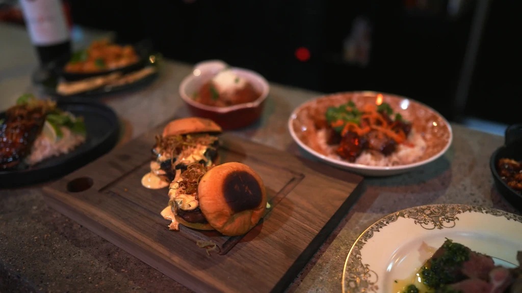 Two gourmet sliders on a wooden board amidst other restaurant dishes.