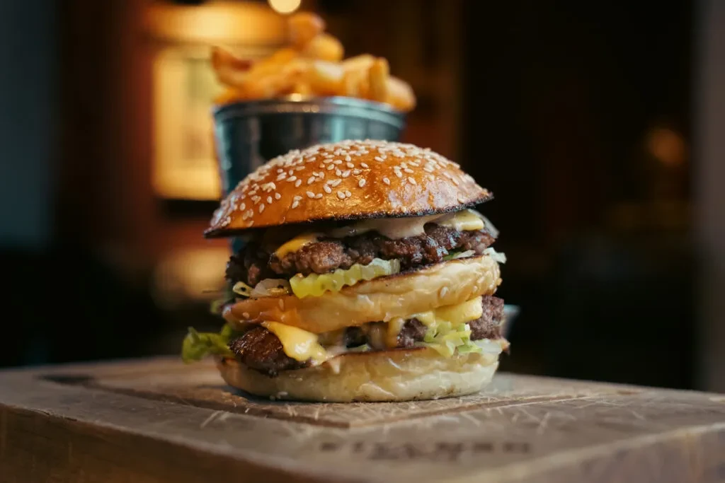 Multi-patty burger with cheese, lettuce on wood, waffle fries behind.