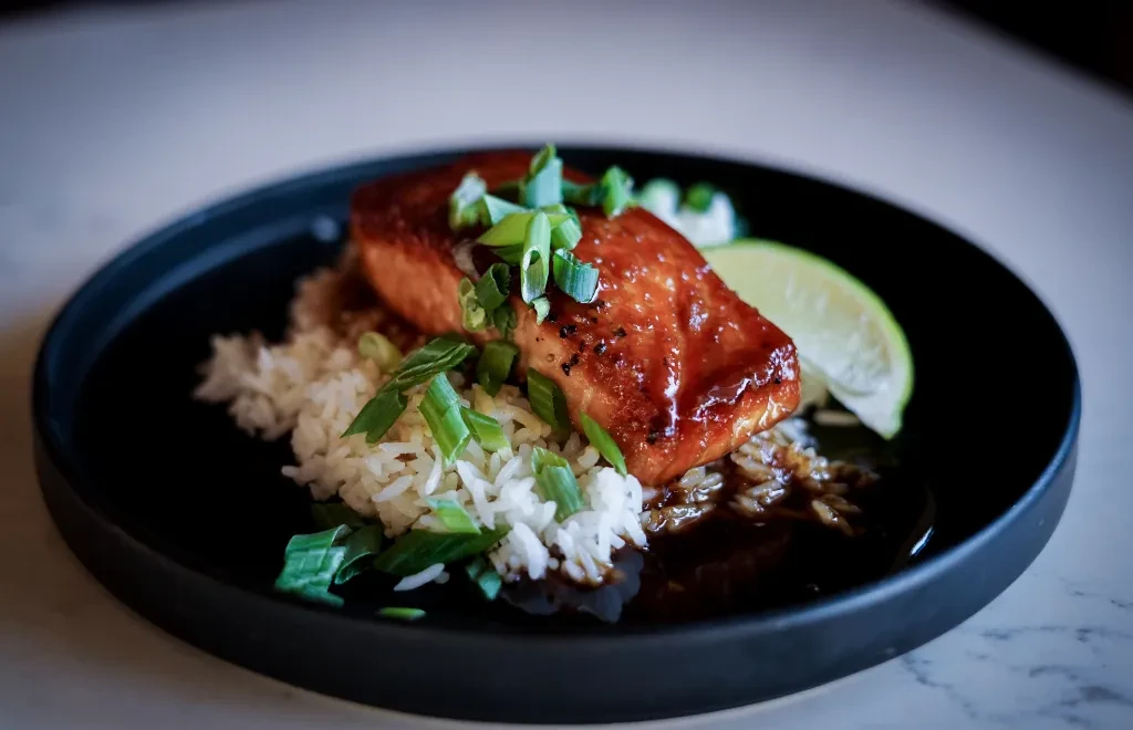 Glazed salmon with rice, green onions, and lime on dark plate.