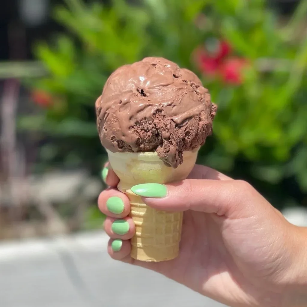 Hand with green nails holding a melting chocolate ice cream cone outdoors.