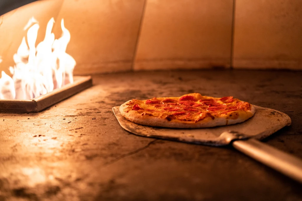 Pepperoni pizza on a peel baking in a fiery stone oven.