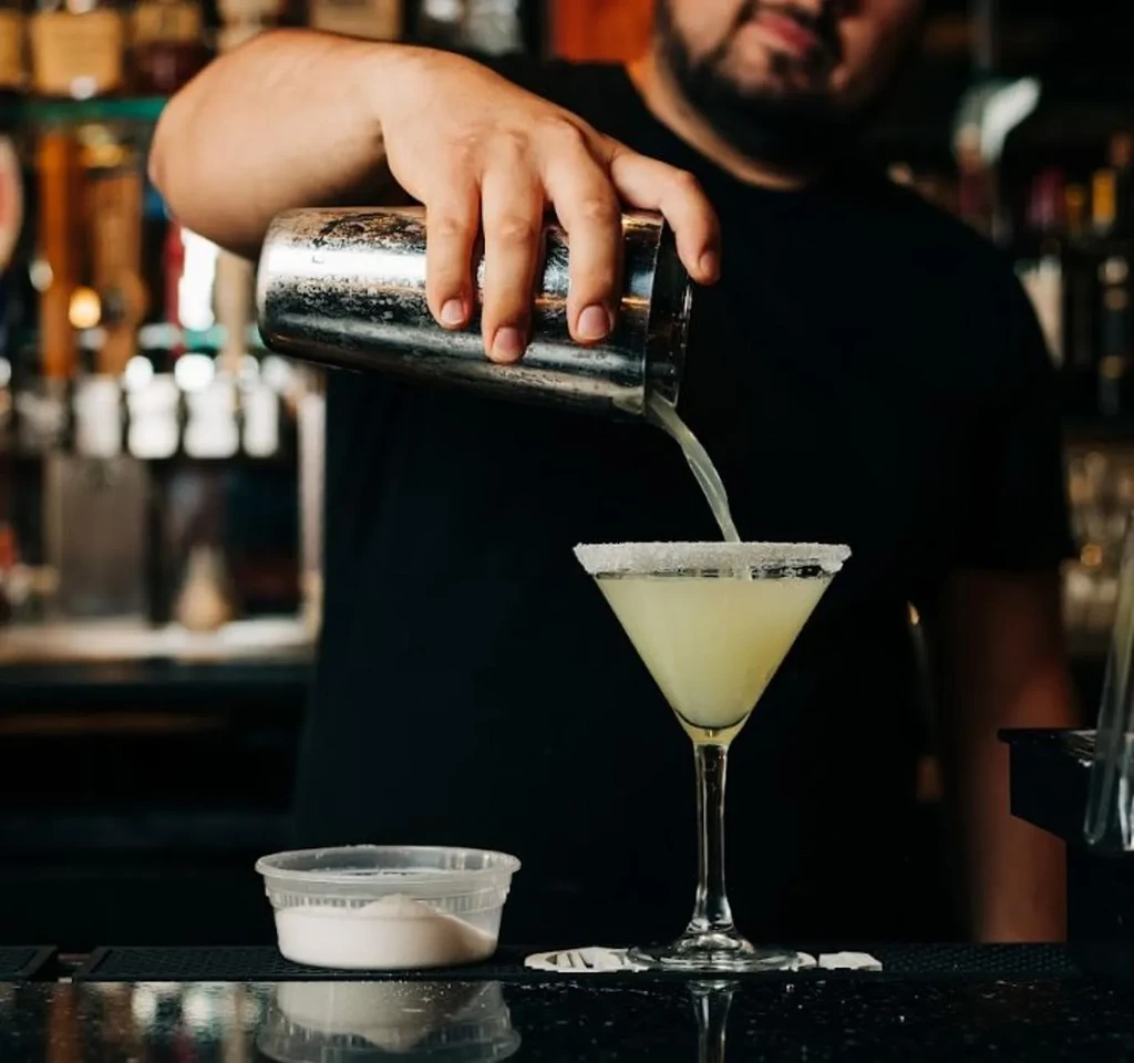 Bearded bartender pours cocktail into a salted rim martini glass.
