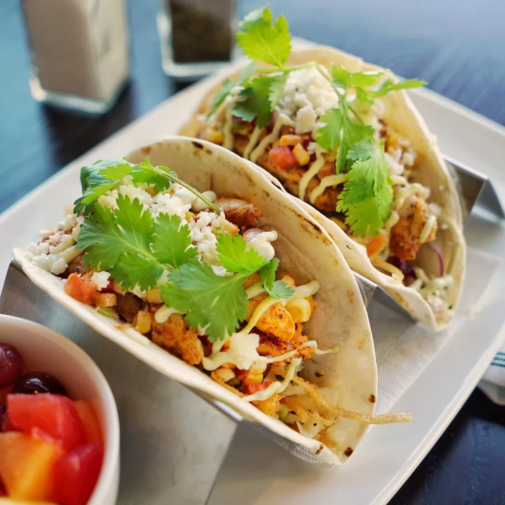 Two colorful tacos in metal stands with a partial fruit salad.
