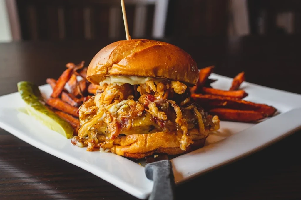 Gourmet burger with melted cheese, sweet potato fries, and pickle spear.