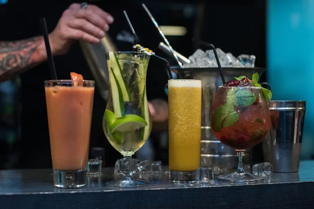 Four colorful, garnished cocktails on a bar; a bartender shakes a drink.