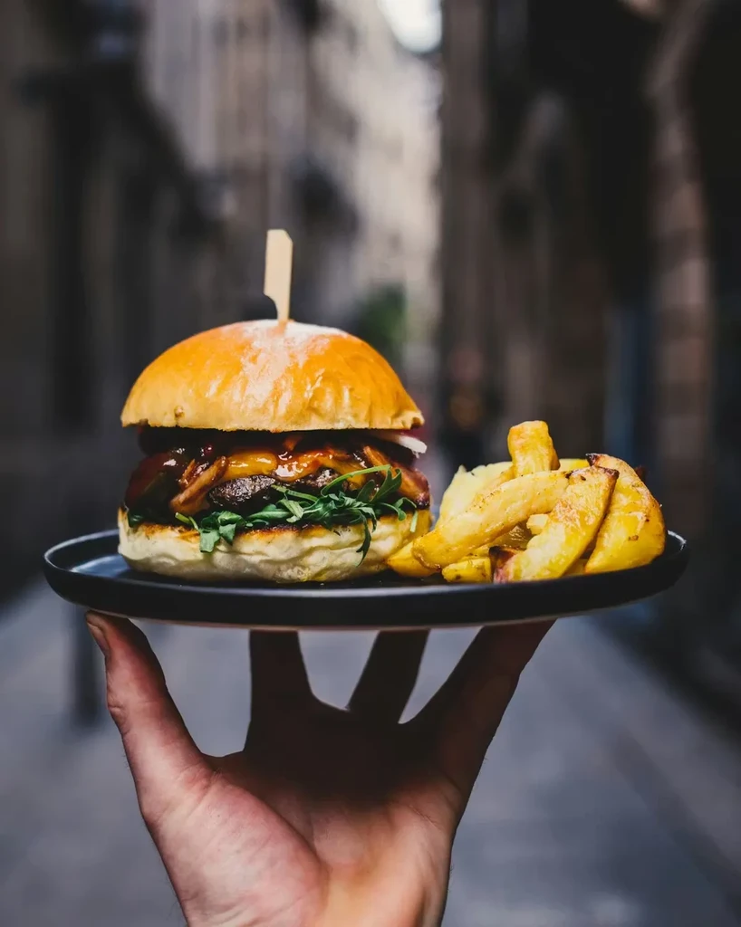 Hand holding gourmet burger with mushrooms and fries, urban background.