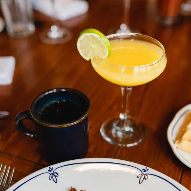 Yellow cocktail, dark mug, and food on a wooden dining table.