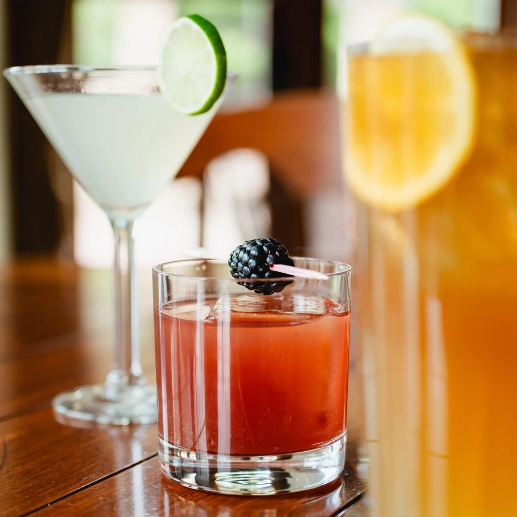 Three drinks on a wooden table: red, clear, and amber, with garnishes.