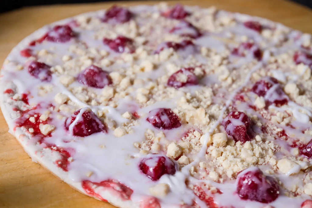 Dessert pizza topped with white frosting, glistening red fruit, and crumbles.