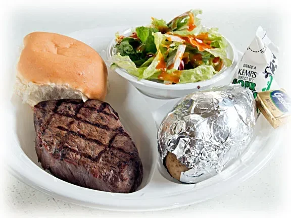 Compartmentalized tray with grilled steak, baked potato, salad, bread, and condiments.