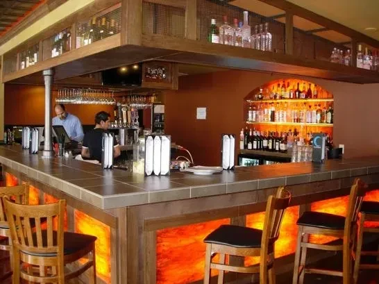 Well-lit L-shaped bar, illuminated orange panels, liquor bottles, two staff.