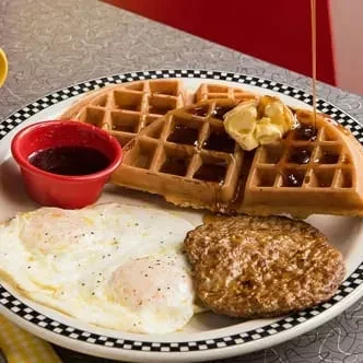 Waffles, fried eggs, sausage, and syrup being poured on a plate.