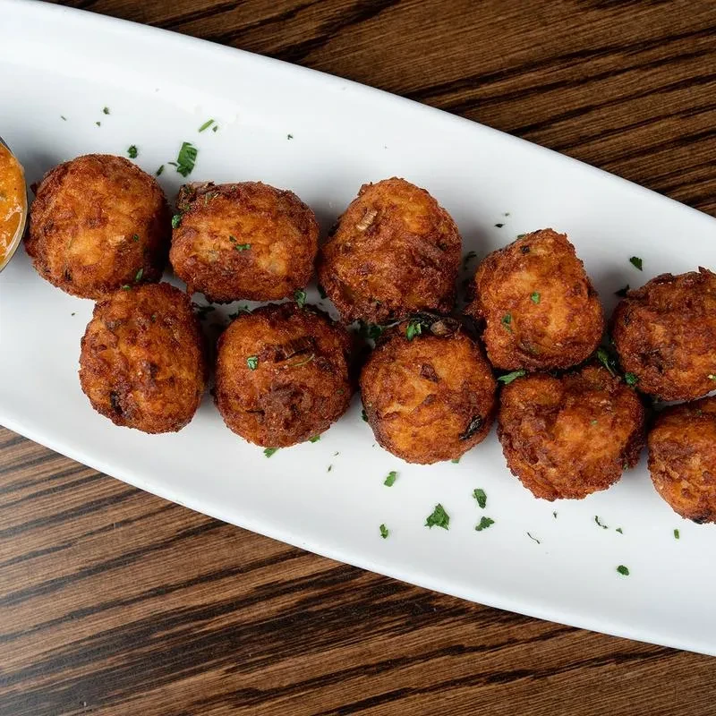 Ten golden-brown fritters with green garnish and orange dipping sauce on wood.