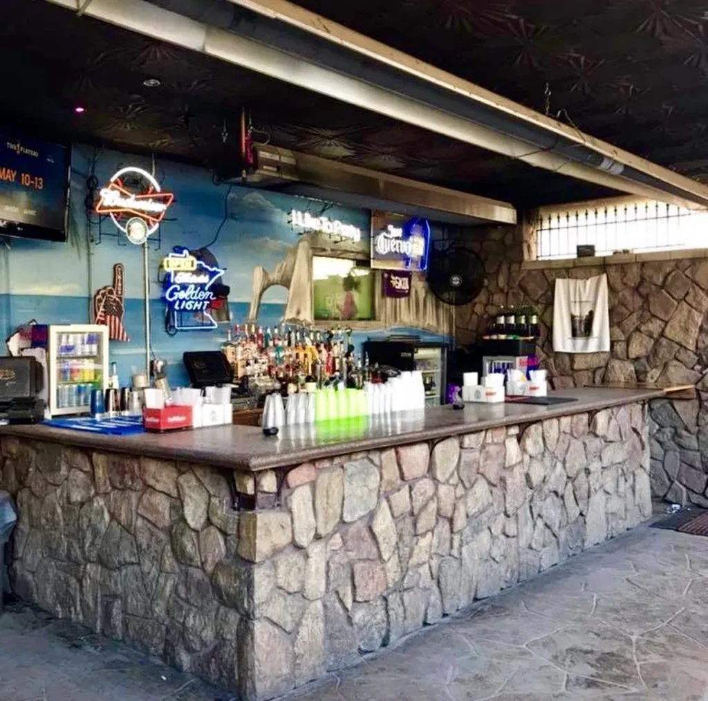 Restaurant bar with stone counter, liquor, coastal mural, and neon signs.