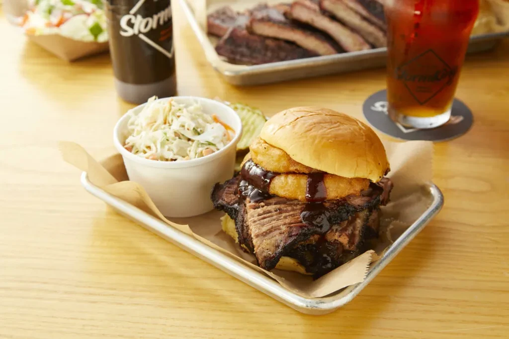 Barbecue brisket sandwich with onion rings, coleslaw, and pickles on a wooden table.