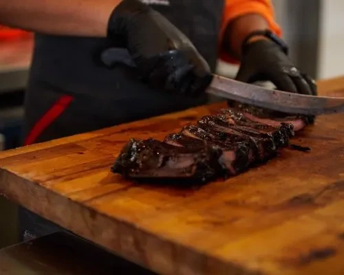 Gloved hands slicing dark, roasted meat on a thick wooden cutting board.