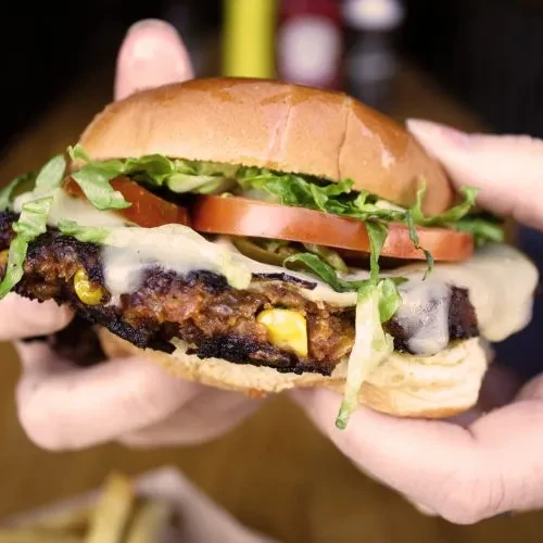 Hands hold a charred burger with corn patty, cheese, and vegetables.