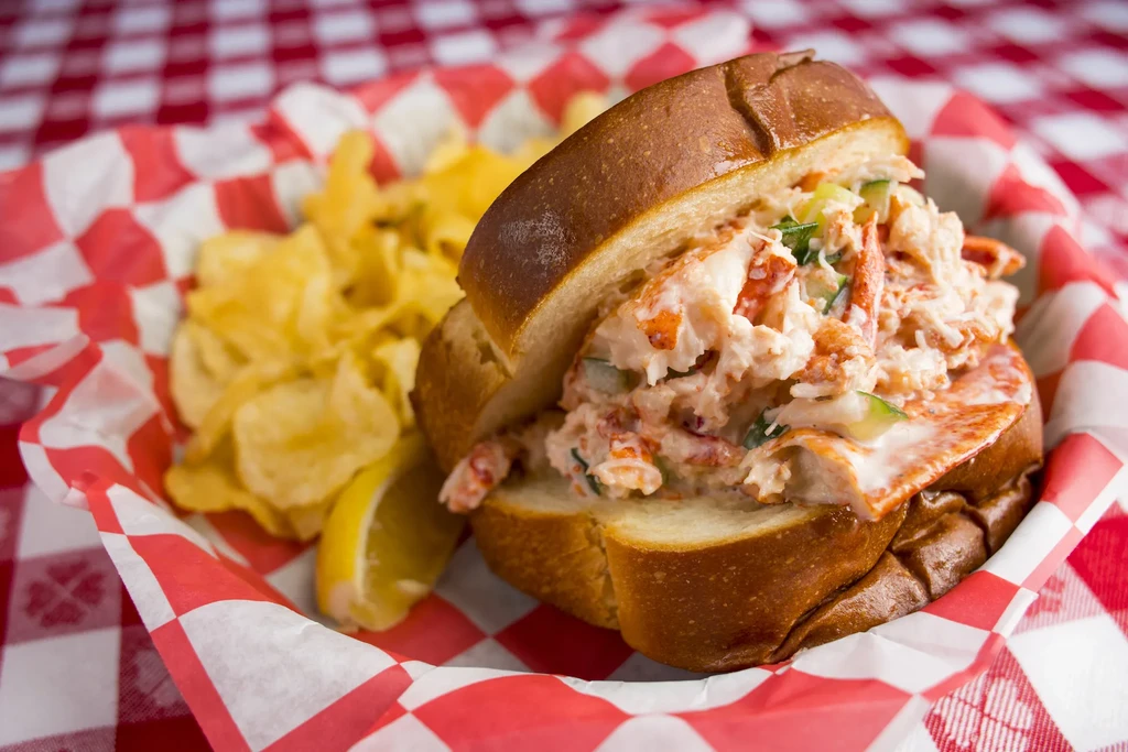 A lobster roll, potato chips, and lemon in a red checkered basket.