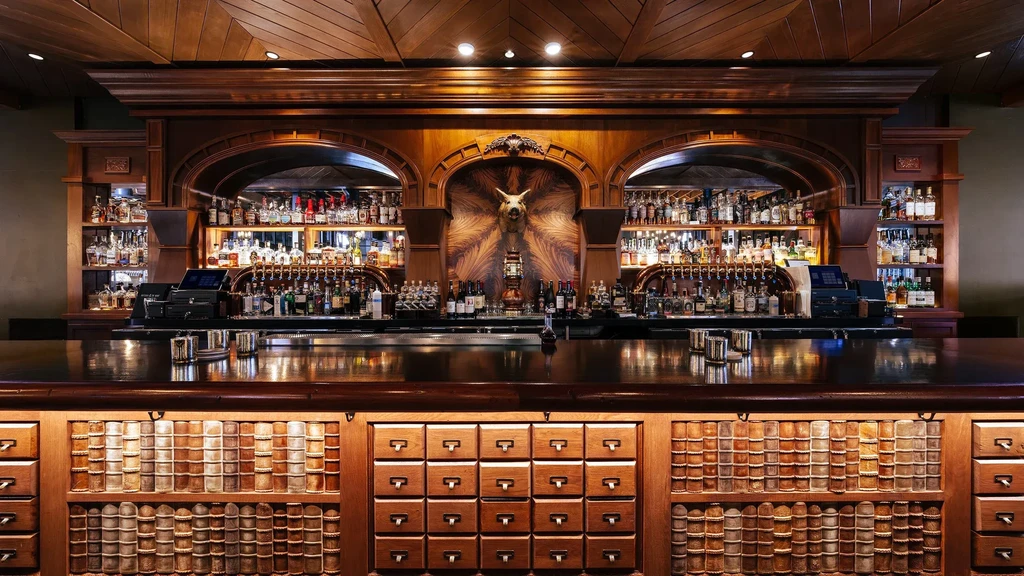 Ornate wooden bar with liquor bottles, taxidermy, and book-paneled front.