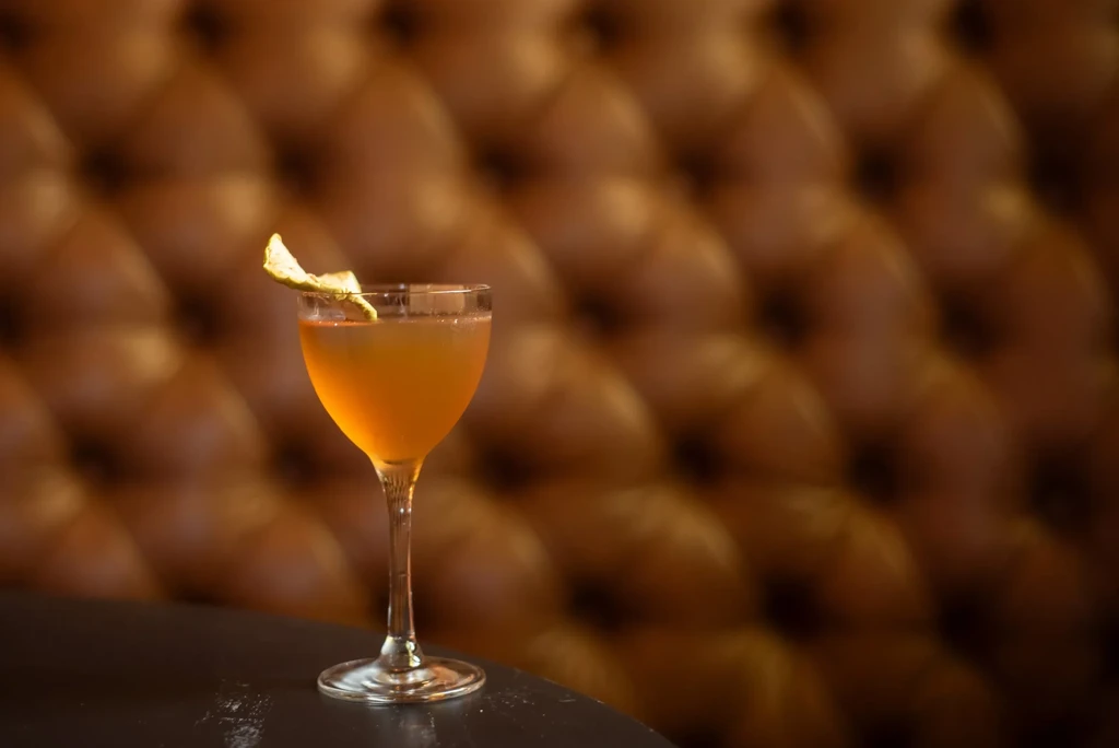 Stemmed amber cocktail with citrus peel against blurred brown upholstery.