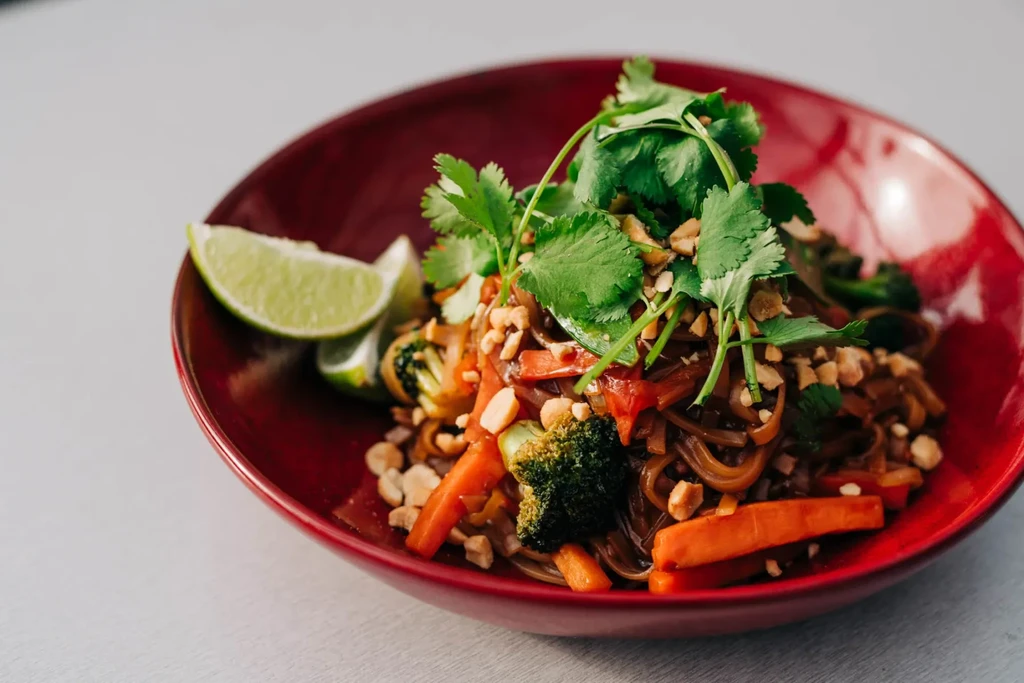 Noodle stir-fry in red bowl with vegetables, cilantro, peanuts, lime.
