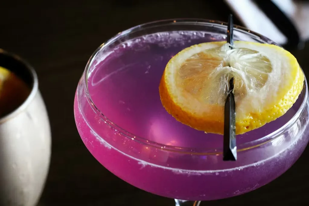 Close-up purple-magenta cocktail in coupe glass with lemon garnish.