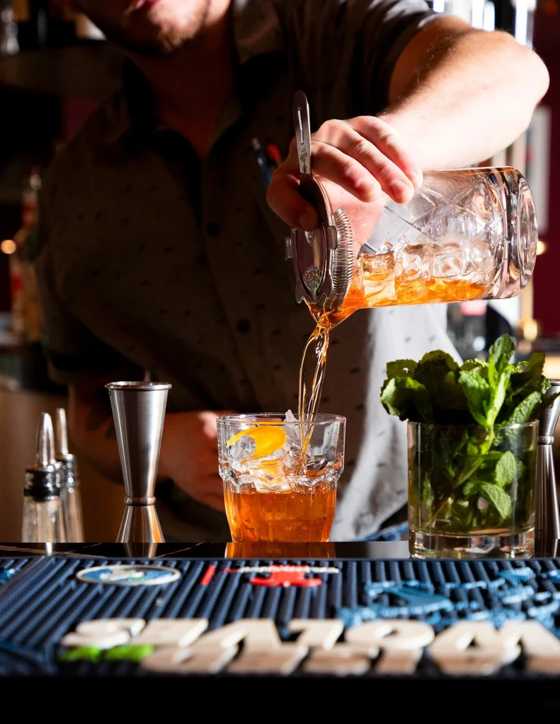 Bartender straining amber cocktail into a rocks glass with ice.