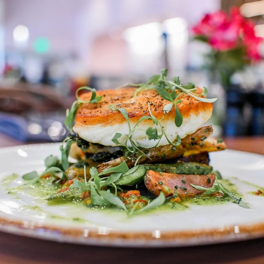 Plated golden-brown seared fish, roasted vegetables, green sauce, microgreens.