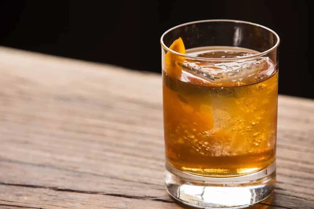 Amber cocktail in clear glass with ice and orange peel on wood.