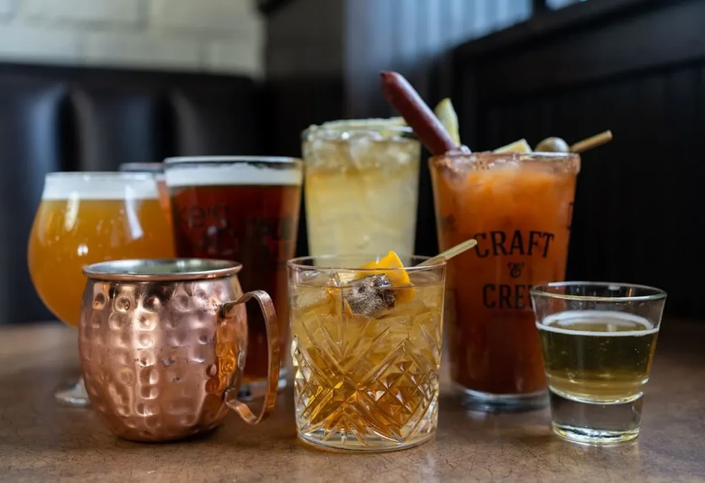 Seven diverse cocktails and beers on a dark bar table.