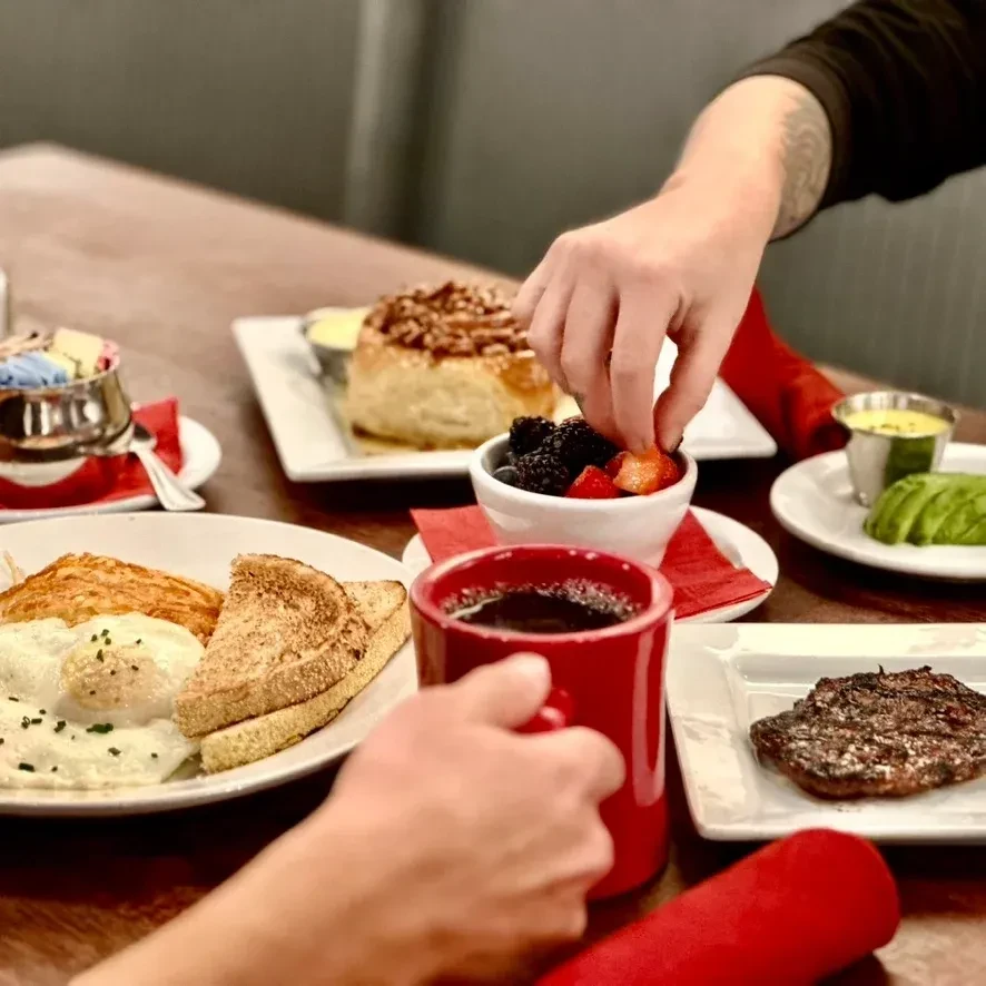 Hands holding coffee and picking berries from a breakfast spread.