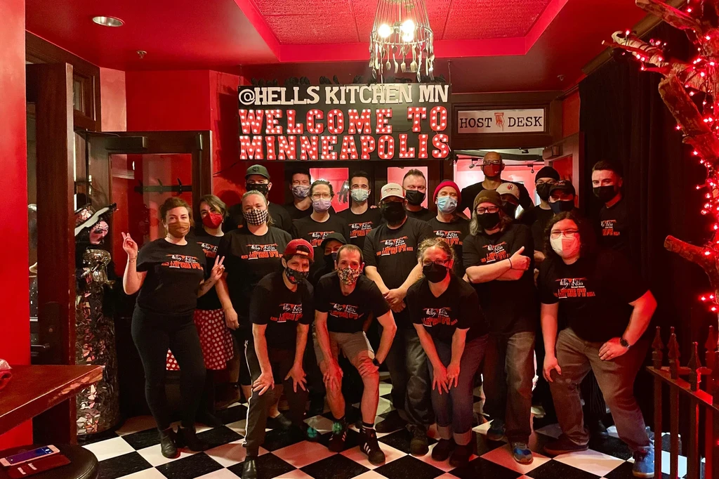 Staff pose under Hells Kitchen MN sign in red restaurant.