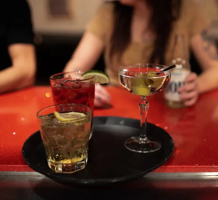 Three distinct cocktails on a bar counter with blurred patrons.