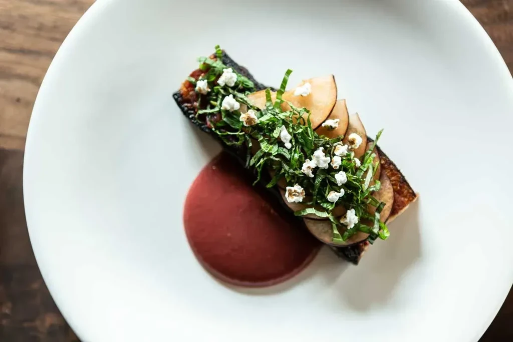 Plated gourmet dish with dark base, green herbs, puffed grains, and purple sauce.