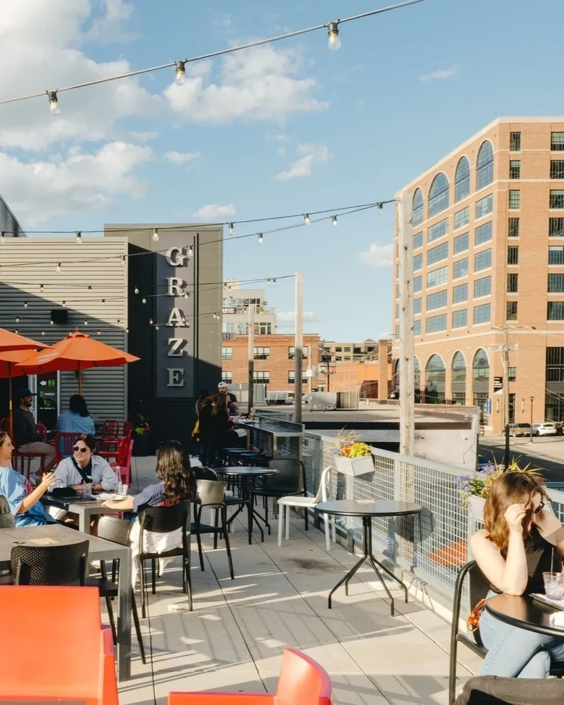 Bustling GRAZE rooftop patio with diners, orange umbrellas, and string lights.