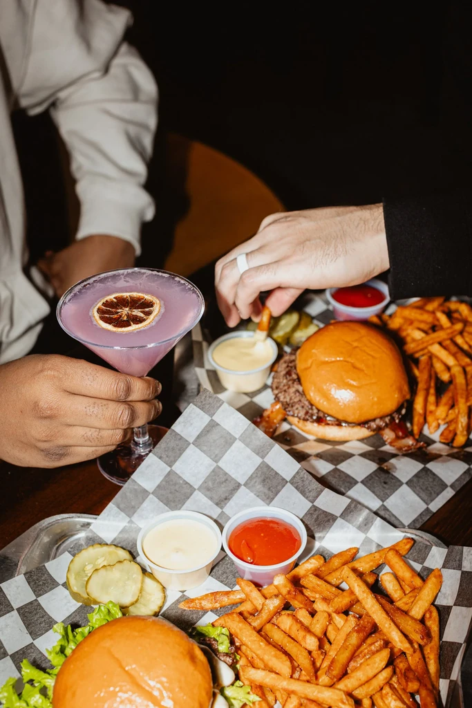Overhead: Two people enjoy burgers, fries, and a purple cocktail.