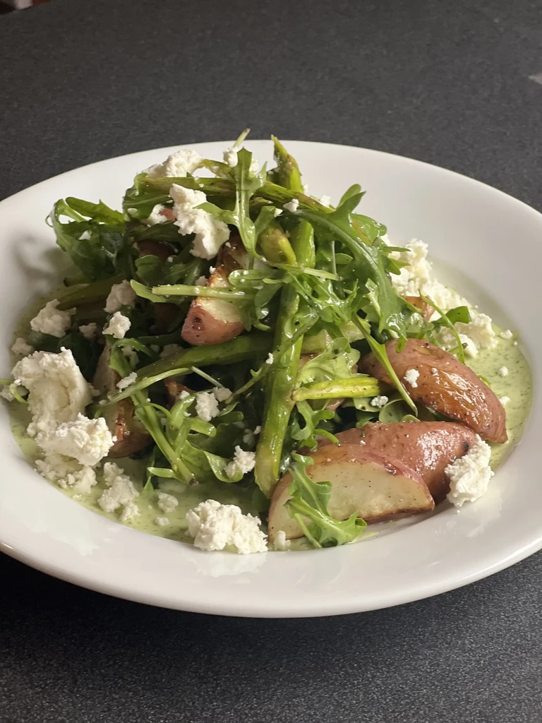 Dish with roasted asparagus, potatoes, arugula, crumbled cheese, green sauce.