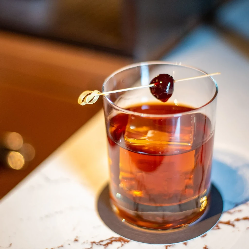 Amber cocktail in rocks glass with large ice cube and cherry.