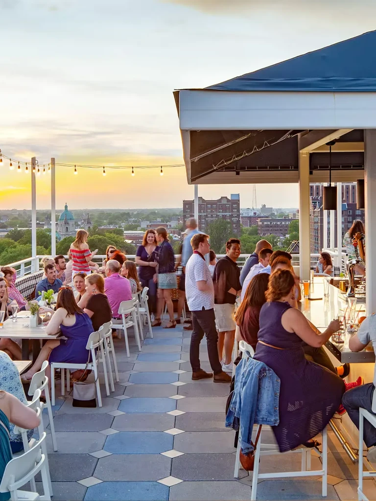 Lively rooftop bar at sunset with patrons, city skyline, and string lights.