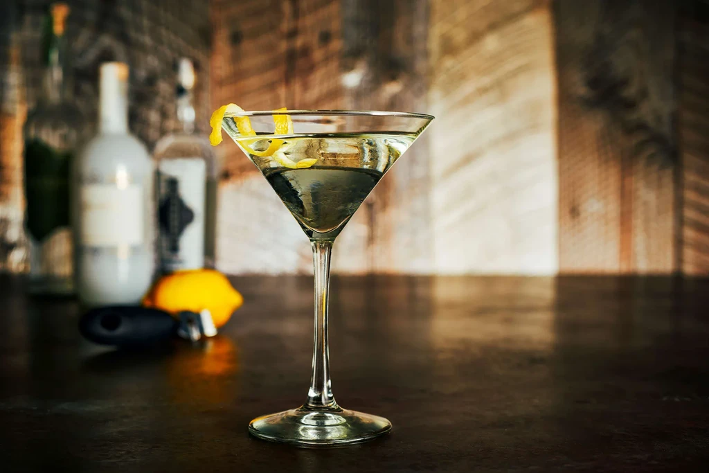 A martini glass with a lemon twist on a bar with bottles.