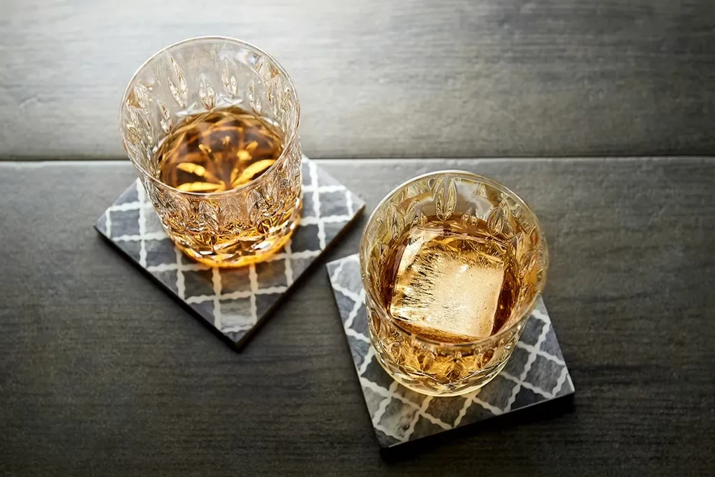 Two crystal glasses, amber liquid, one ice cube, on patterned coasters.