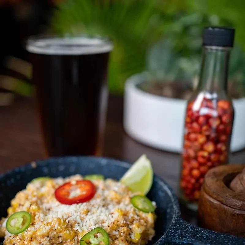 Esquites in dark bowl with cheese, jalapeños, and a lime wedge.