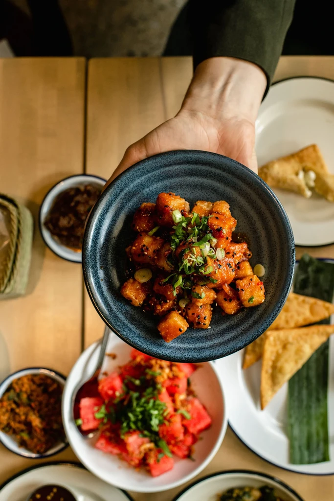 Top-down photo: hand holding sauced food bowl, surrounded by other dishes.