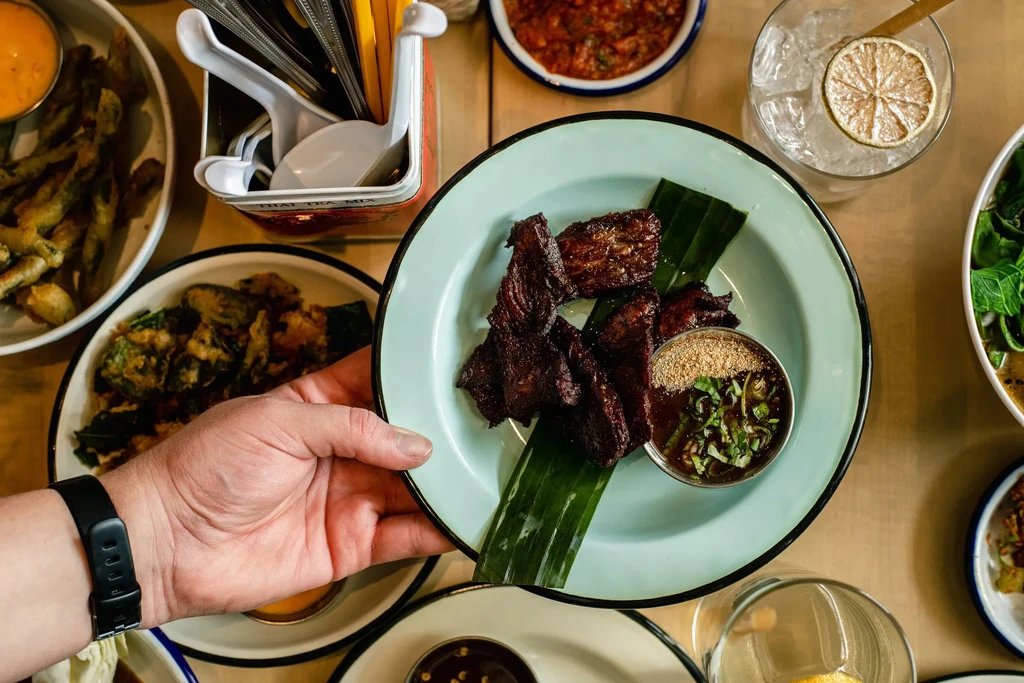 Overhead: Hand holds blue plate of meat with banana leaf, sauce.