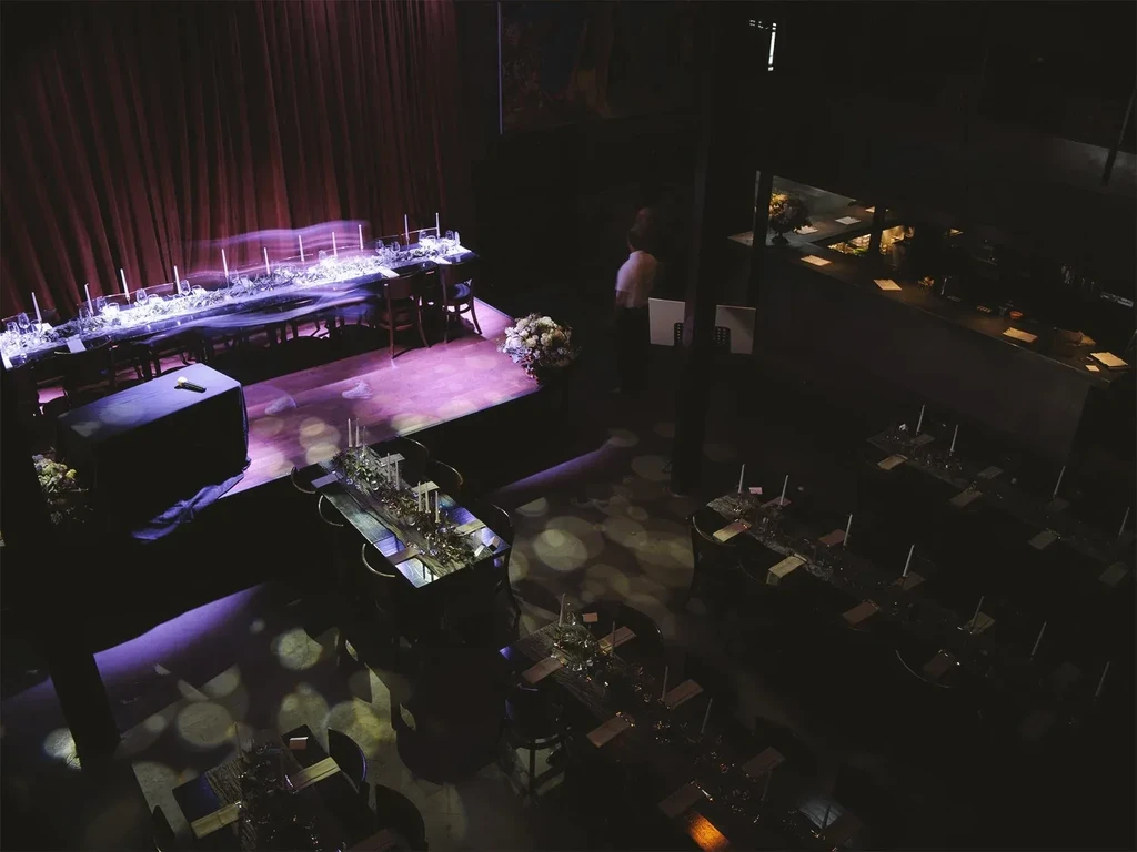 Dimly lit event venue with stage, elegantly set tables, purple lights.