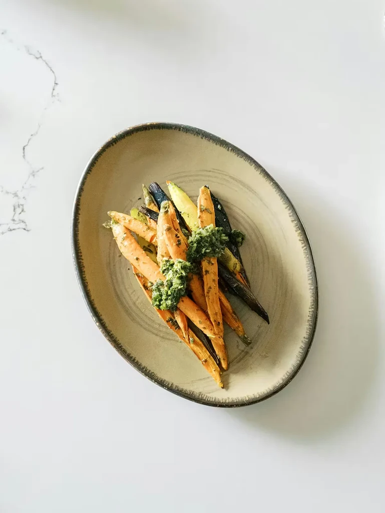 Roasted orange and purple carrots with green herb sauce on a plate.