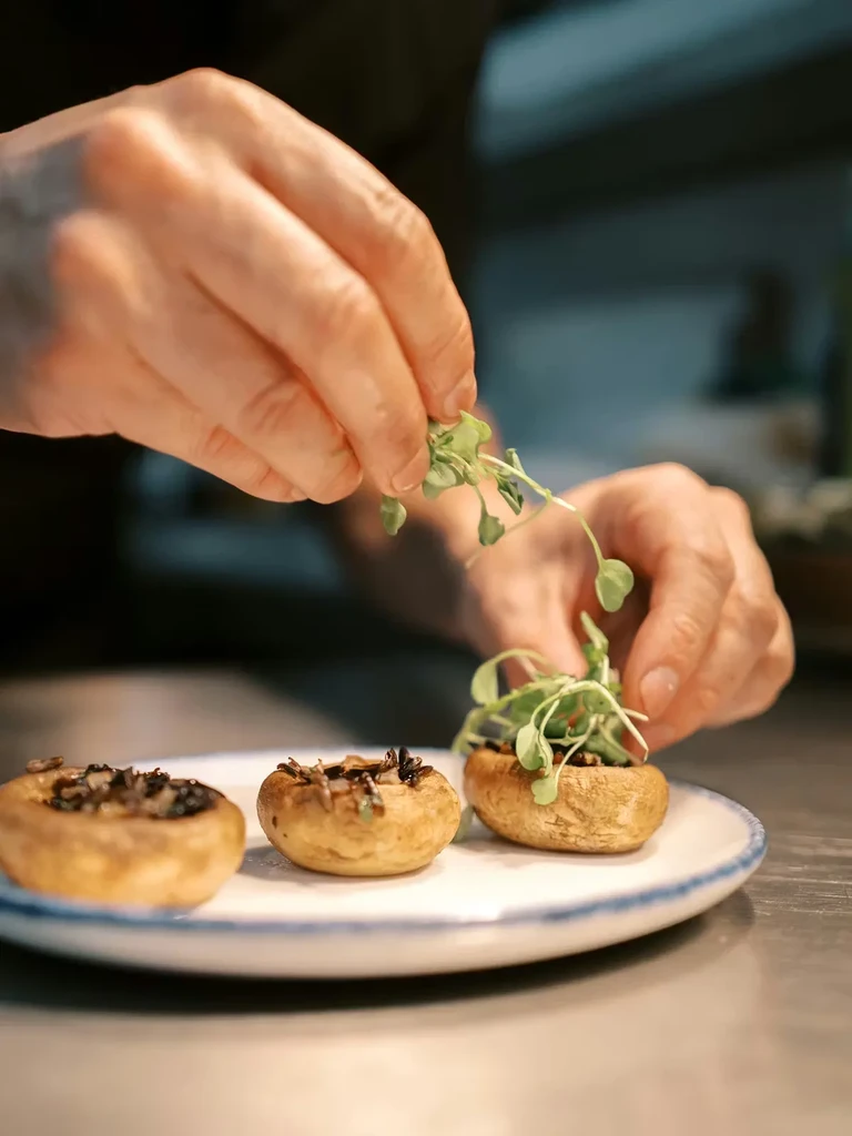 Hands garnish three stuffed mushrooms with fresh green microgreens.