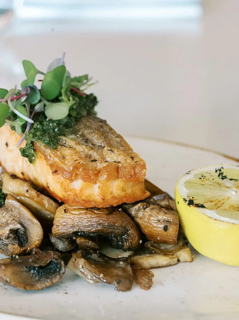 Close-up of pan-seared salmon, mushrooms, green sauce, and charred lemon.